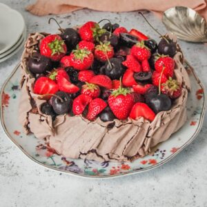 A chocolate pavlova with strawberries and cherries sits on a floral ceramic plat on a light gray surface.