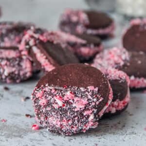 A chocolate peppermint sandwich cookie decorated with crushed candy canes rests on another cookie on a light gray surface.