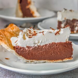 A slice of chocolate pudding pie sits on an individual white ceramic plate on a gray surface.