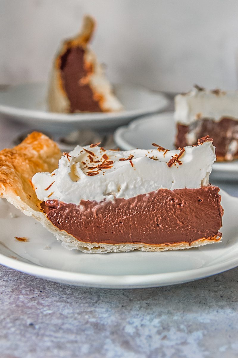 A slice of chocolate pudding pie sits on an individual white ceramic plate on a gray surface.