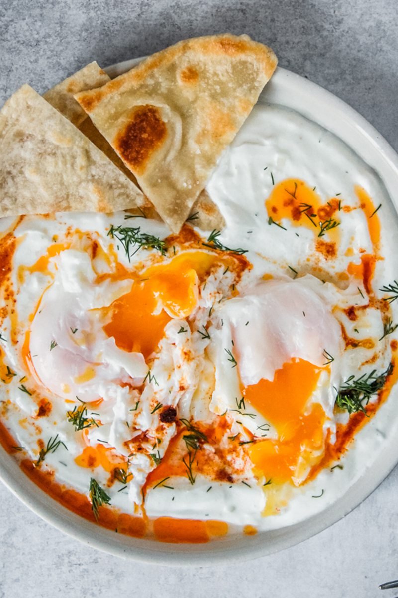 Cilbir - Turkish Poached Eggs in Yogurt sit broken in a rimmed ceramic plate with a few pieces of bread served on the side on a gray surface.