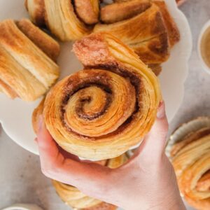 A German cinnamon roll is held up with it's layers of pastry visible above a larger plate of cinnamon rolls sitting below on a gray surface.