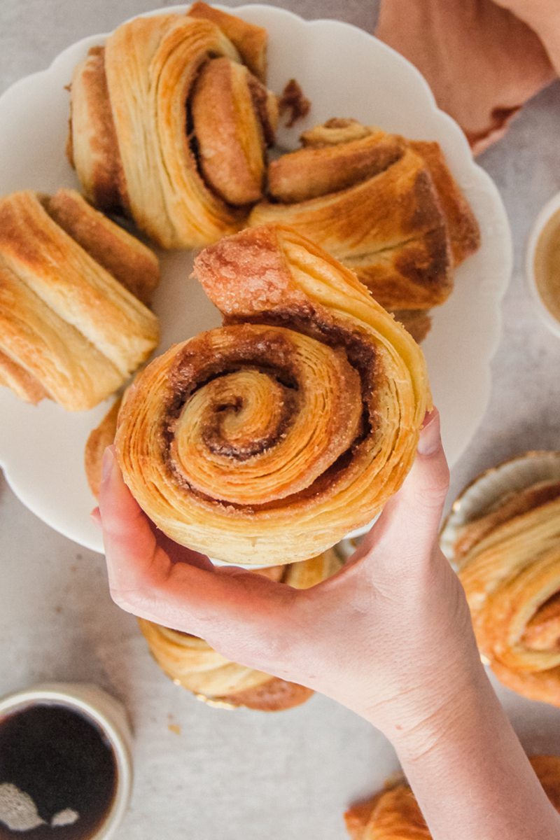 A German cinnamon roll is held up with it's layers of pastry visible above a larger plate of cinnamon rolls sitting below on a gray surface.