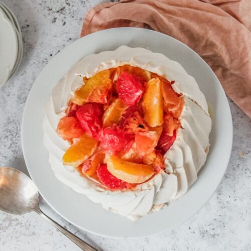 Oven roasted citrus sits atop a pavlova served on a white plate on a light gray surface.