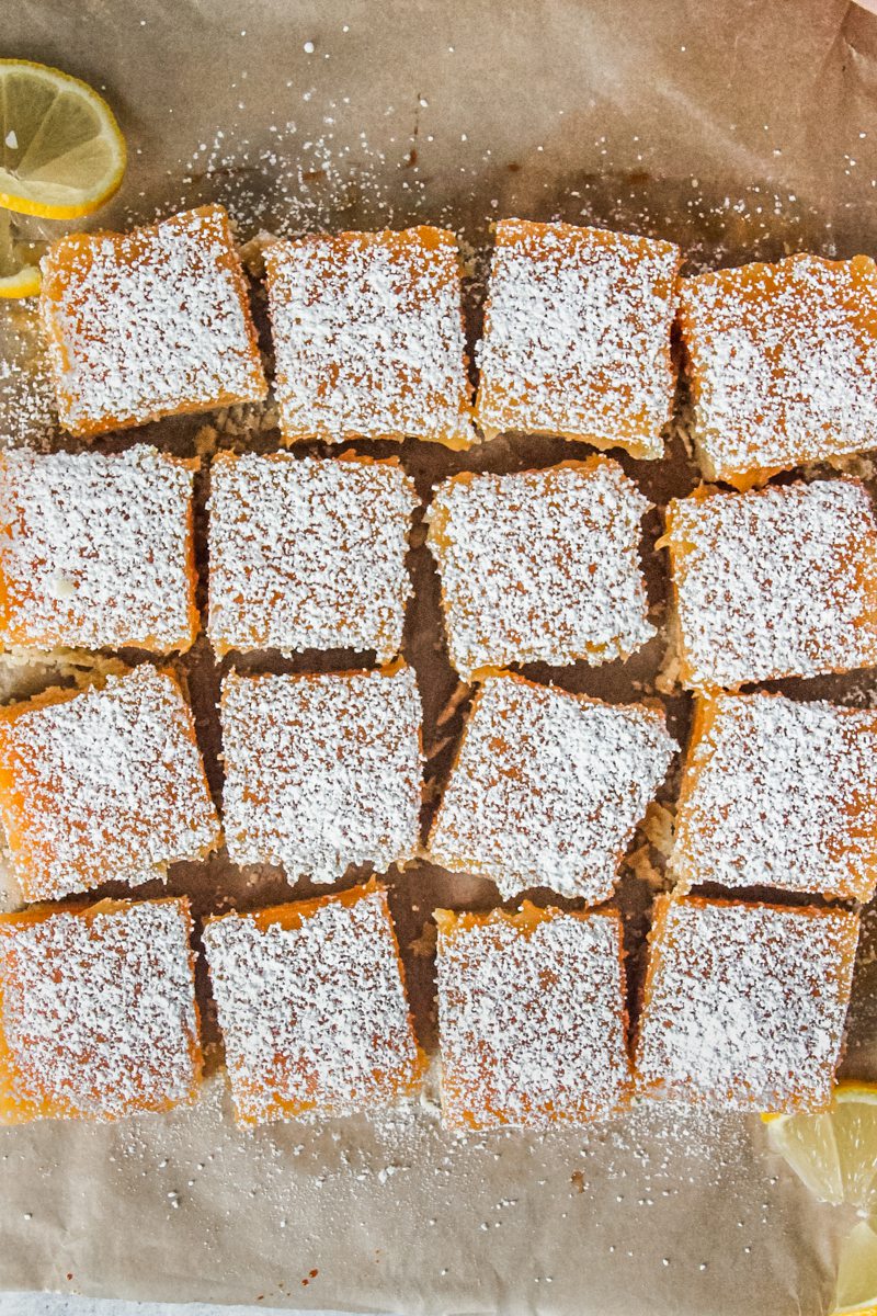 Squares of lemon curd bars topped with a sprinkling of powdered sugar on top sit on a brown parchment paper surface.