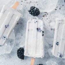 Coconut Blackberry Popsicles lay on top of ice cubes on a light grey surface.