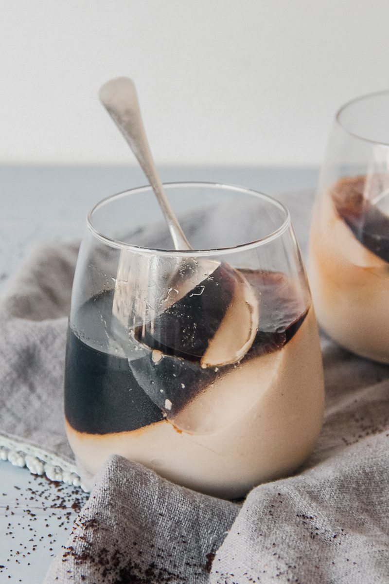 A spoonful of panna cotta rests on the remaining dessert in a glass on a gray cloth on a blue gray surface.