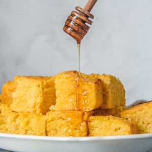 Honey is drizzled over squares of apple cornbread sitting on a white ceramic plate on a gray surface.