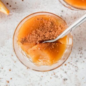 An individual serving of Costa Rican Orange Pudding topped with ground cinnamon sits on a light gray surface with a spoon tucked in.