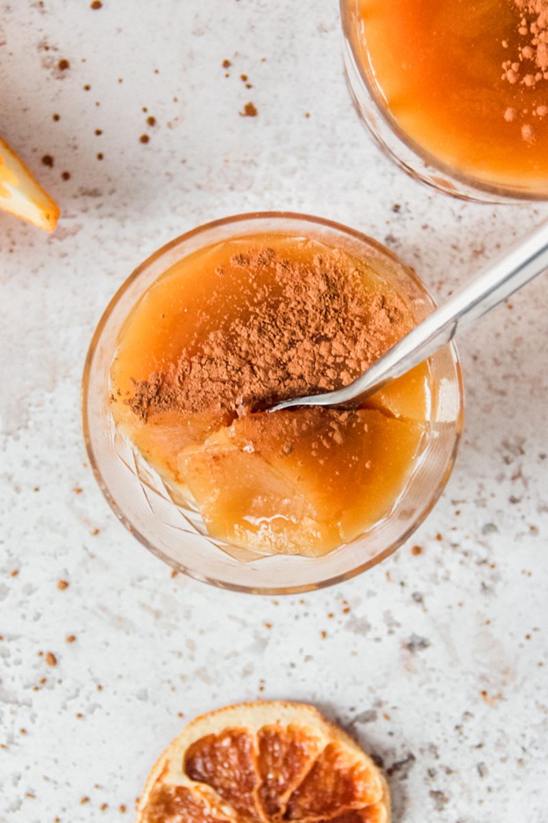 An individual serving of Costa Rican Orange Pudding topped with ground cinnamon sits on a light gray surface with a spoon tucked in.