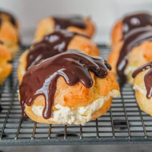 A close up of a cream puff halved wtih a whipped cream filling and topped with a melted chocolate topping sits on a cooling rack above a gray surface.