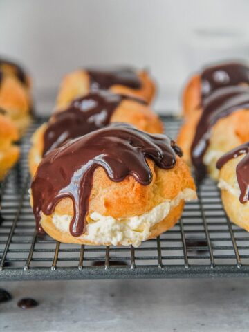 A close up of a cream puff halved wtih a whipped cream filling and topped with a melted chocolate topping sits on a cooling rack above a gray surface.