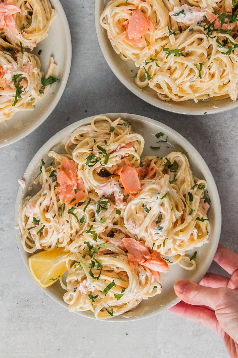 A plate of creamy smoked salmon pasta topped with finely chopped parsley and a lemon wedge on the side is held above a gray surface.