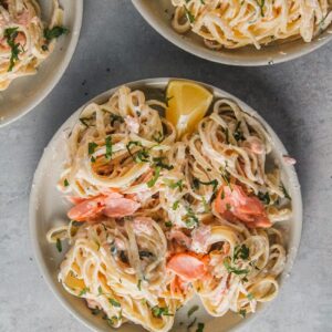 A rimmed gray ceramic plate with creamy smoked salmon pasta topped with finely chopped parsley and a lemon wedge sits on a gray surface.