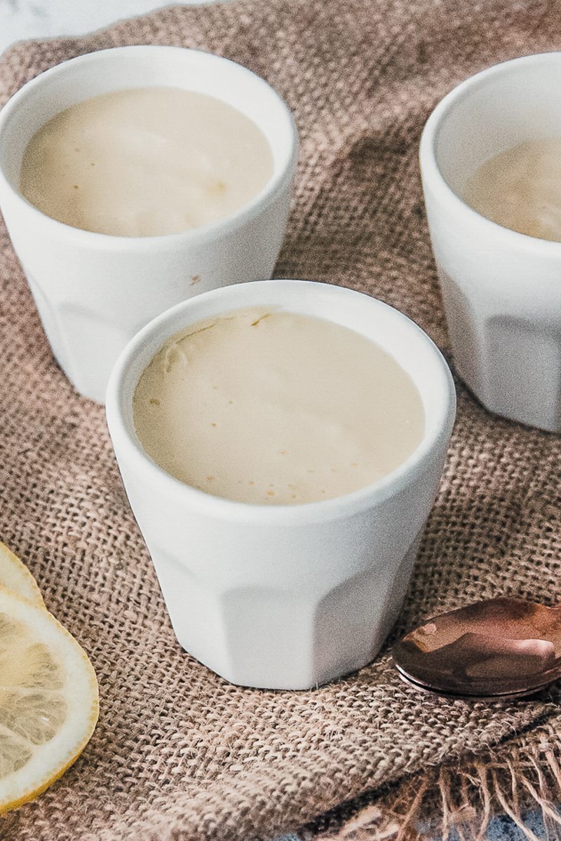 Lemon posset sits in white ceramic espresso cups on brown cloth.