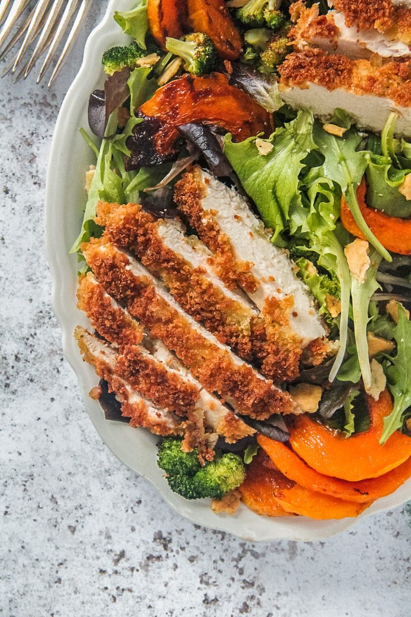 A close up of breaded crispy chicken slices on top of lettuce and roasted butternut squash served up on a white plate on a gray surface.