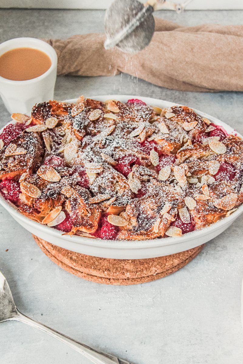 Powdered sugar is sprinkled on top of croissant bread pudding in an oval white baking dish above a gray surface.