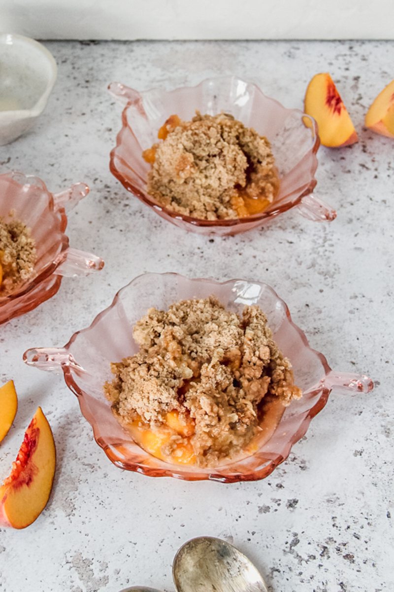 Peach crumble is served in individual pink glass bowls on a light gray surface.