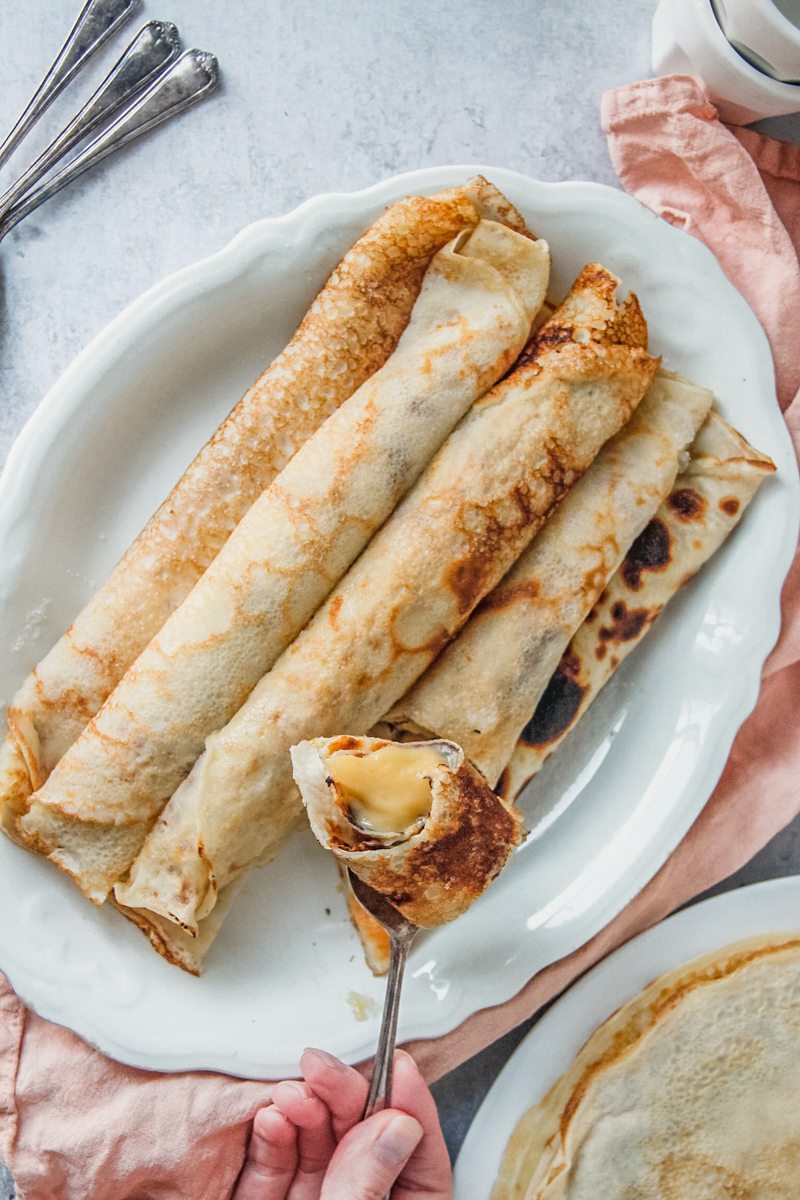 A spoonful of rolled pancakes filled with custard is held above a plate of rolled filled pancakes on an oval white ceramic plate on a gray surface.