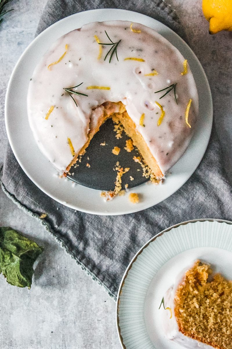 A lemon rosemary cake sits on a white ceramic plate with a slice cut out sitting on an individual plate beside.