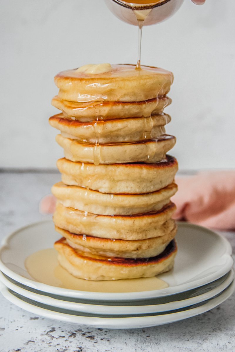 Honey is poured over a stack of griddle cakes sitting on a stack of white ceramic plates on a light gray surface.