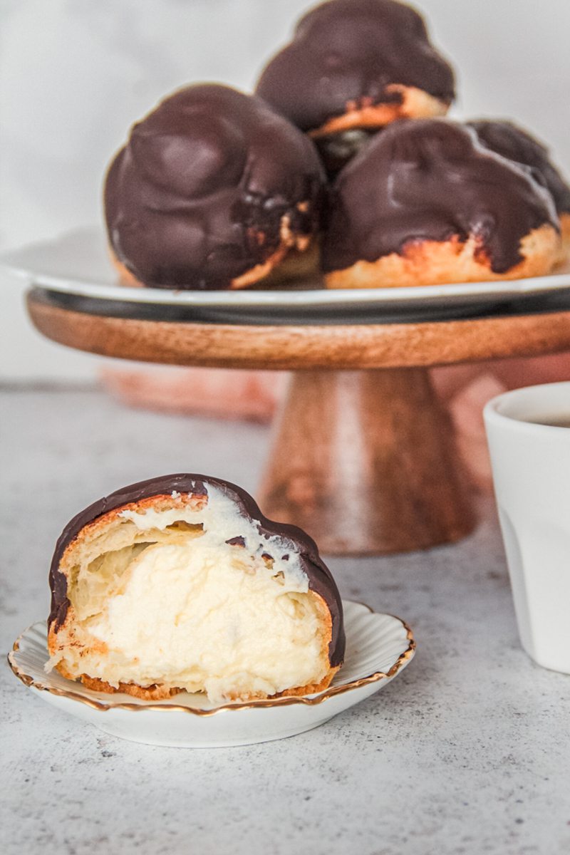 A halved Dutch profiterole sits on an individual plate filled with whipped cream and with a chocolate topping and cake stand on a light gray surface.