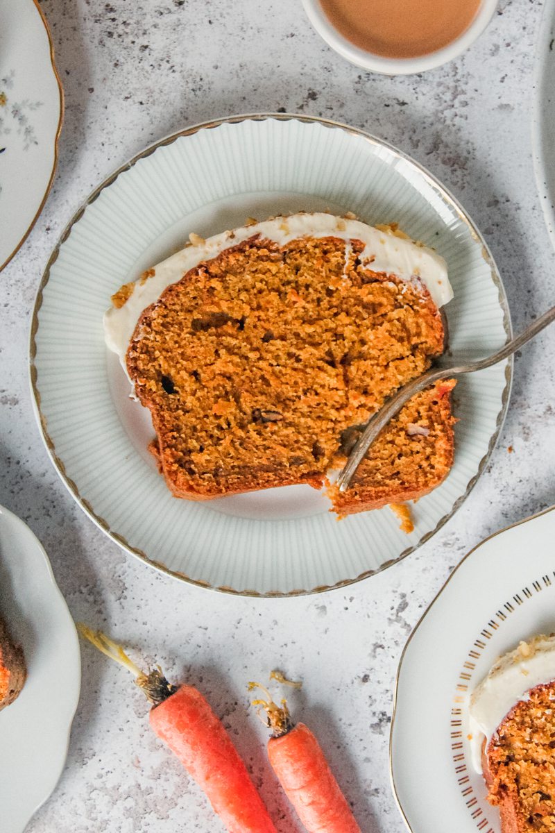 A slice of carrot cake loaf with a corner cut off by a dessert fork resting on the edge of an individual ceramic plate on a light gray surface.