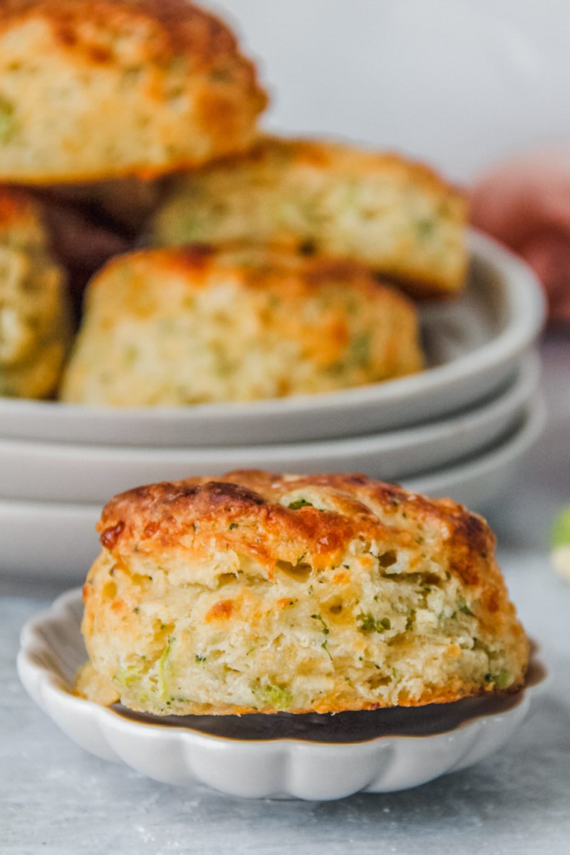 A savory cheddar cheese biscuit sits on a small ceramic plate with a batch sitting on rimmed ceramic plates on a gray surface.