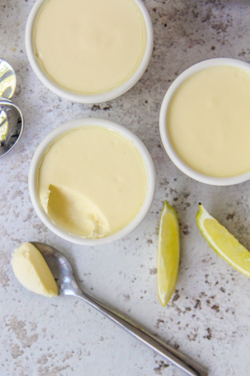 Lemon posset in white ceramic espresso cups with a spoonful on the side and a couple of lemon wedges and spoons beside on a light gray surface.