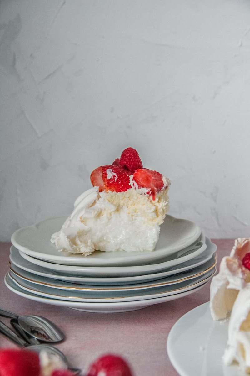 A serving of pavlova at a side angle on a stack of plates with the marshmallow interior visible topped with whipped lemon cream and fresh berries.