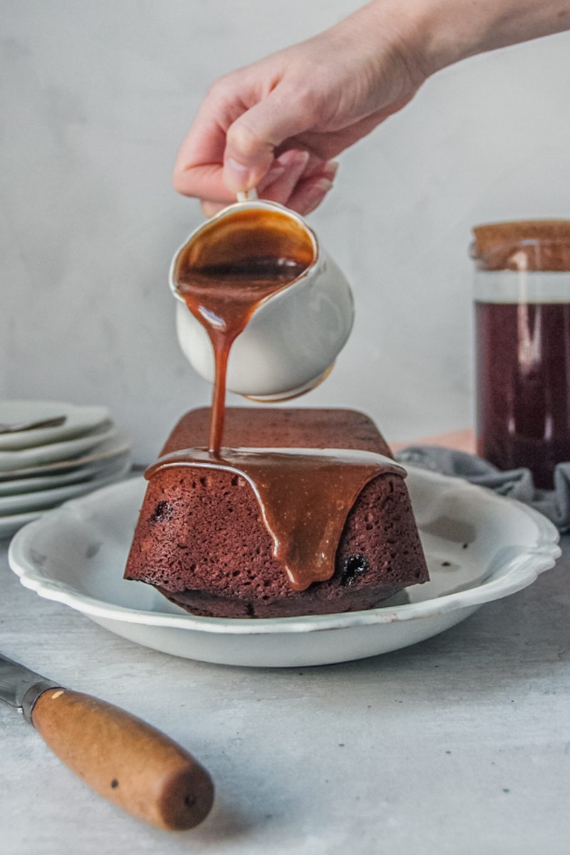 A sticky toffee pudding loaf cake is topped with toffee sauce on a white oval plate on a gray surface.