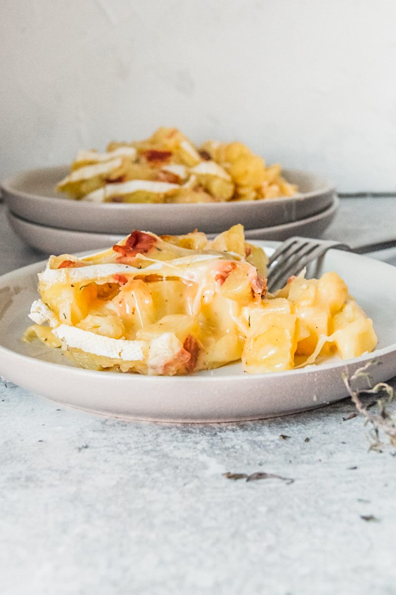 An individual serving of Tartiflette sits on a rimmed ceramic plate on a light gray surface.