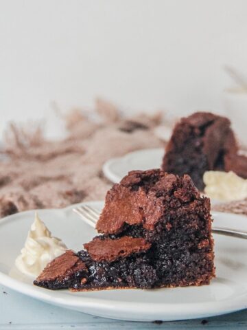 A slice of Fudgy Swedish Chocolate cake is served up on an individual white ceramic plate with it's gooey interior visible beside a dollop of whipped cream on a light blue background.