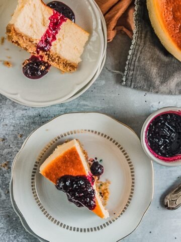 Two slices of New York Cheesecake sit on individual plates topped with a berry sauce on a gray surface.