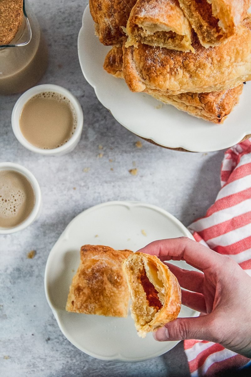 A halved Sintra Pillow is held up with it's flaky pastry enveloping an almond filling with other sintra pillows served up on a plate beside and small white ceramic cups of tea.