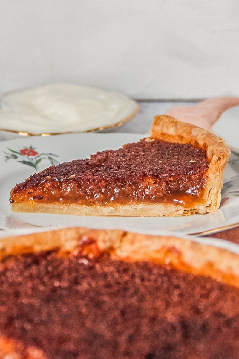 A single slice of treacle tart with it's gooey center and crispy pastry sits on an individual ceramic plate on a light gray surface.