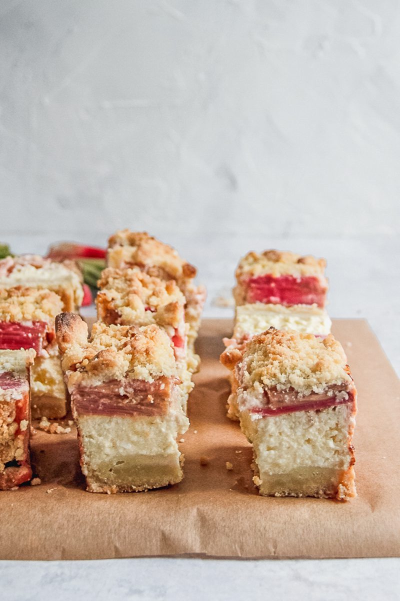 Squares of Estonian Rhubarb Custard and Streusel Cheesecake sit on parchment paper on a gray surface.