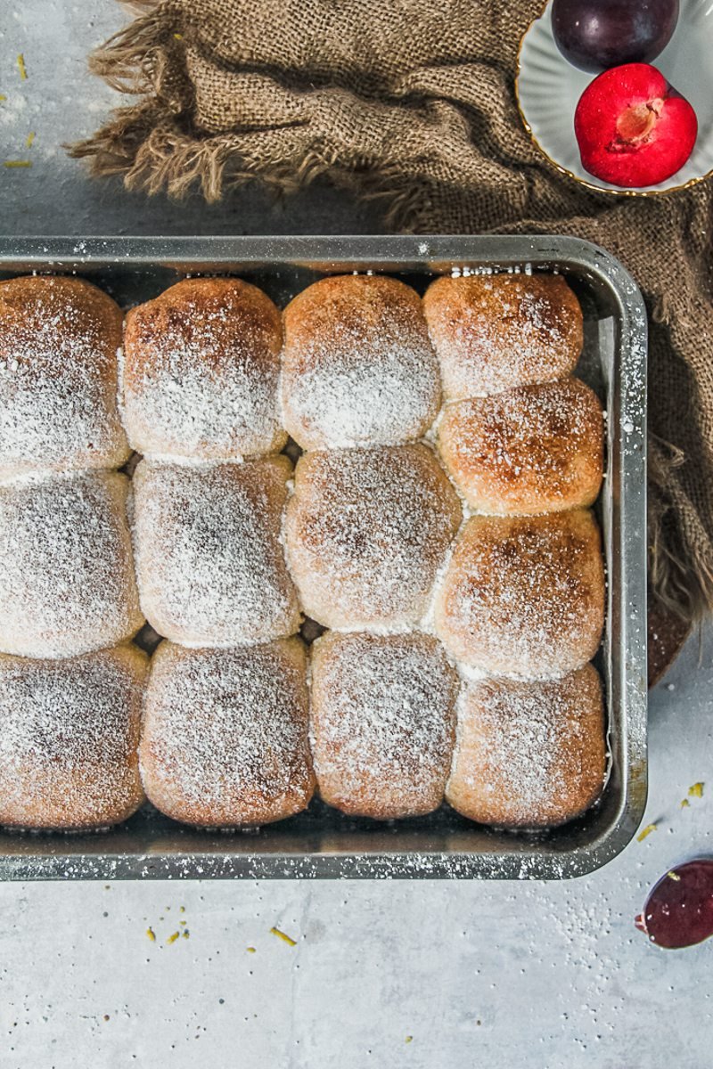 Jam filled rolls sit in a stainles steel baking tin beside a brown cloth on a gray surface.