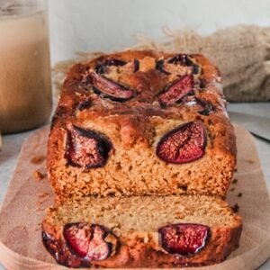 A slice of fig loaf cake with baked fresh figs on top sits on it's side with the warm brown interior visible on a wooden board on a gray surface.