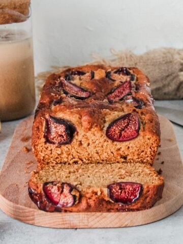 A slice of fig loaf cake with baked fresh figs on top sits on it's side with the warm brown interior visible on a wooden board on a gray surface.