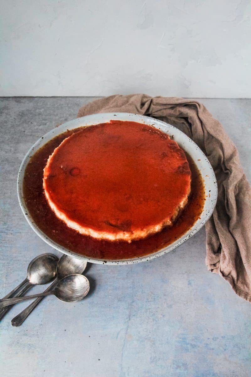 A flan dessert is served up on a ceramic blue plate sitting on a gray surface.