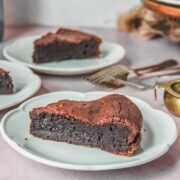 A slice of French chocolate cake sits on an individual white ceramic plate with it's fudgy center visible on a light pink background.