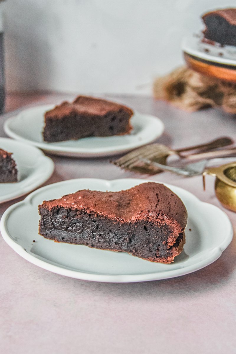 A slice of French chocolate cake sits on an individual white ceramic plate with it's fudgy center visible on a light pink background.