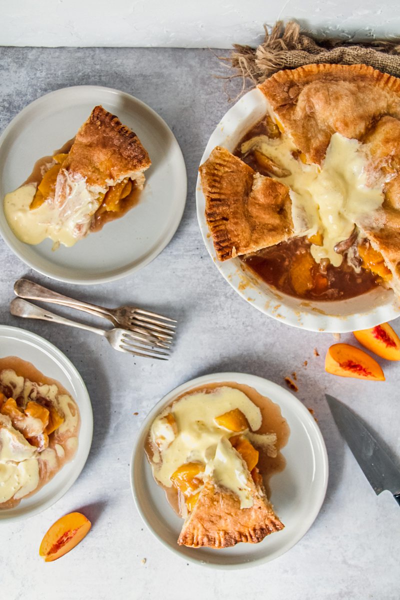 Individual servings of double crusted peach cobbler sit on plates with melting vanilla ice cream mixed with the peach juices beside the remaining pie on a gray surface.