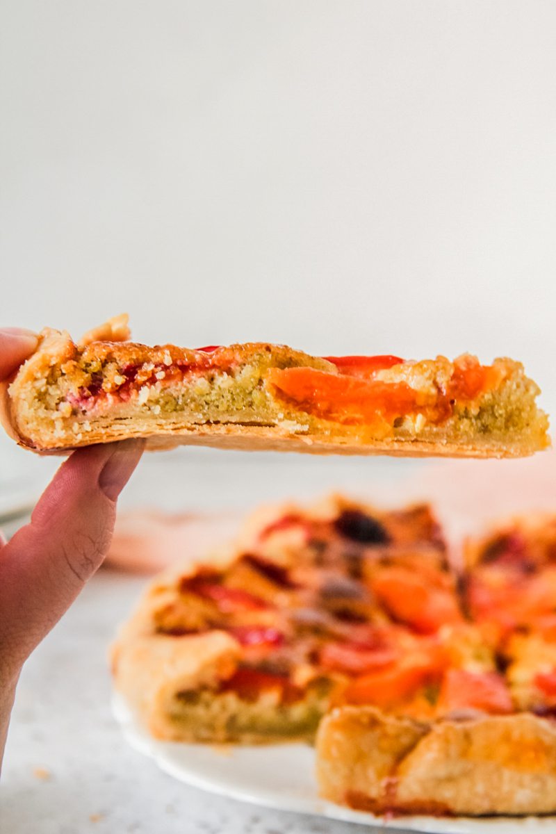 A slice of apricot galette with a pistachio frangipane filling is held up to show off the layers in front of the remaining galette.