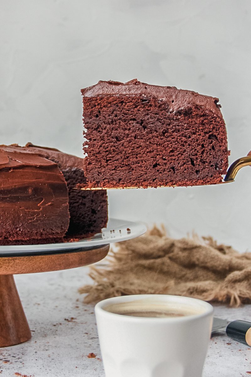 A slice of chocolate buttermilk cake sits on a cake server above a light gray surface with a fork piercing the front of the cake.