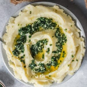 A side angle of mashed potatoes topped with a garlic herb butter mixture on a white plate on a gray surface beside a brown cloth.