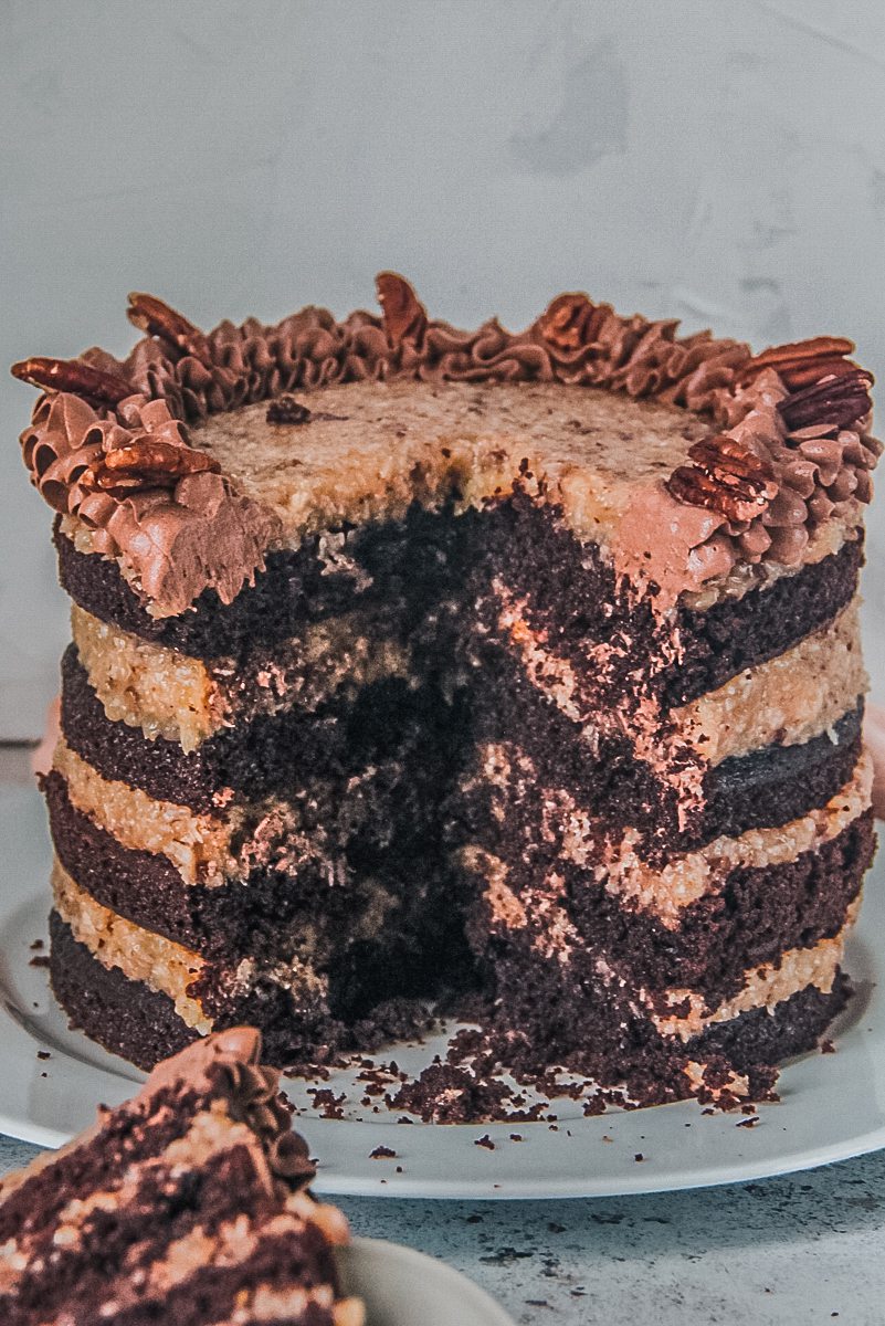 Layers of German chocolate cake with a pecan coconut filling sit on a white ceramic plate on a light gray surface.