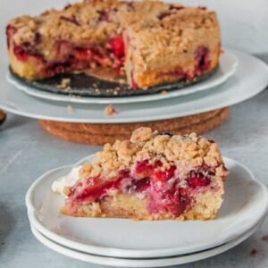 A slice of plum cake sits on two white plates with the remaining cake on white plates on a gray surface.