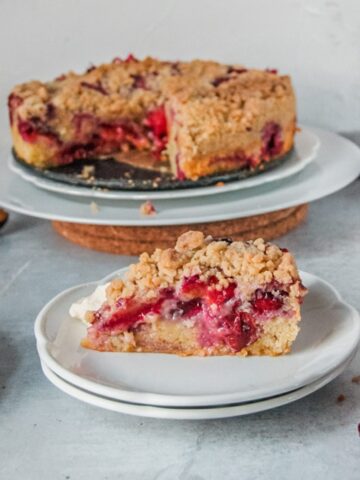 A slice of plum cake sits on two white plates with the remaining cake on white plates on a gray surface.